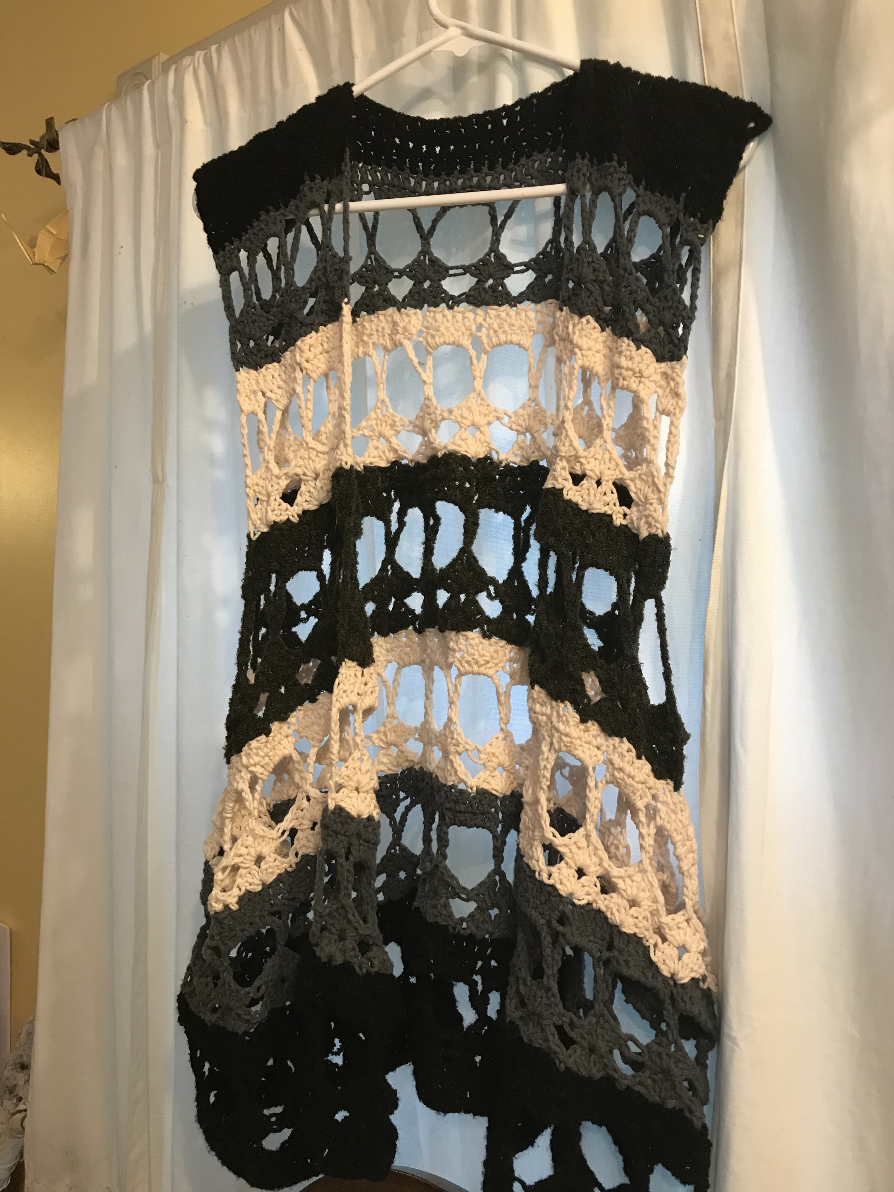 Photo of a crocheted vest hanging in front of a white window curtain. The vest has one central dark green stripe surrounded by two stripes each of white, gray, and black: the colors, more or less, of the agender pride flag